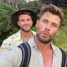 two men standing next to each other in front of some trees and bushes with one man taking a selfie