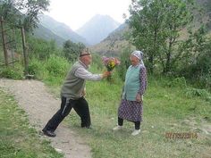 an older couple is standing in the grass with flowers and looking at each other's hand