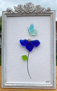 a blue and green glass flower in a silver frame on a wooden table next to a white wall