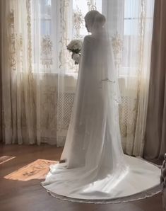 a woman in a white wedding dress standing by a window with her veil pulled back