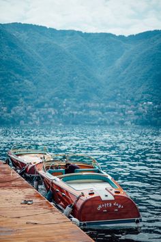 two boats are docked at the end of a wooden pier on a lake with mountains in the background