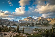 a lake surrounded by mountains and trees under a cloudy blue sky with clouds in the background