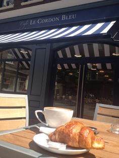 a croissant and cup on a table in front of a restaurant with striped awnings