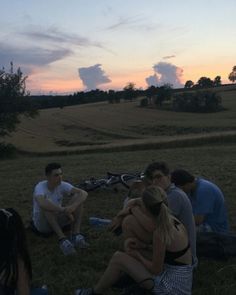 group of people sitting on the grass in front of an open field with trees at sunset