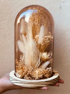 a hand holding a glass dome with dried flowers and feathers in it