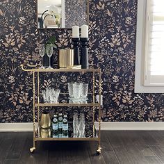 a gold bar cart with glasses and bottles on it in front of a floral wallpaper