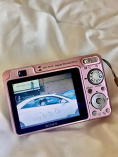 a pink digital camera sitting on top of a bed next to a remote control device