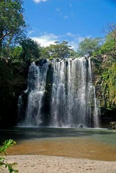 a large waterfall in the middle of a forest with clear blue skies above it and brown water below