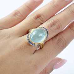 This exquisite ring features a stunning oval-shaped Aquamarine at its center, surrounded by a delicate halo of Iolite accent stones. The soft blue hue of the Aquamarine contrasts beautifully with the deep violet of the Iolite, creating a harmonious blend of colors. The ring is crafted from luxurious yellow gold, enhancing the brilliance of the gemstones. The intricate lattice work on the shank adds a touch of elegance and sophistication, making this piece a perfect statement accessory for any oc Luxury Oval Topaz Ring With Gemstone Accents, Oval Sapphire Ring With Gemstone Accents, Elegant Oval Cabochon Topaz Ring, Exquisite Oval Gemstones With Accents, Blue Art Deco Gemstone Rings, Elegant Oval Multi-stone Topaz Ring, Oval Multi-stone Sapphire And Topaz Ring, Elegant Oval Halo Ring With Multi-stone, Art Deco Aquamarine Ring Vintage