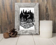 a white frame with a black and white drawing of a cabin in the woods next to a candle