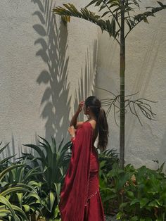 a woman in a red dress standing next to a tree and some green plants on the side of a building