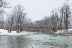 a wooden bridge over a river surrounded by snow covered trees and bare trees in the background