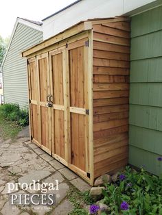 a wooden storage shed sitting next to a green house with purple flowers in the front yard