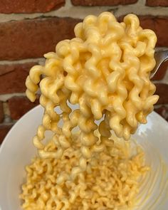 a spoon full of macaroni and cheese being held over a white plate with red brick wall in the background