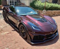 a maroon sports car parked in front of a house on a brick driveway next to a stone wall