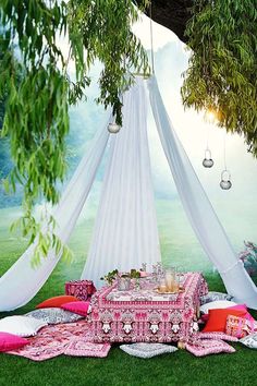 a pink and white table setting under a tree in the grass with hanging decorations on it