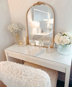 a white vanity with a mirror, stool and flowers on it's side table