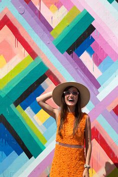 a woman in an orange dress and hat standing next to a colorful wall