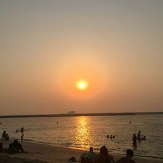 people are sitting on the beach as the sun sets in the distance over the water