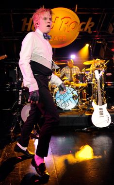 a man with pink hair and white shirt standing on stage in front of musical instruments