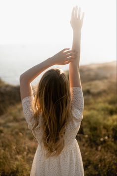 a woman in a white dress standing on top of a grass covered field with her hands up