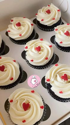 twelve cupcakes with white frosting and red hearts on them in a box