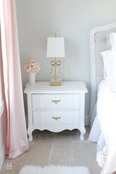 a white nightstand sitting next to a bed with pink curtains on the window sill