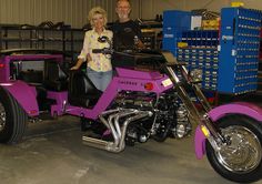 a man and woman standing next to a pink motorcycle in a garage with boxes on the wall