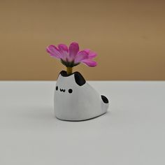 a white vase with a black and white cat holding a pink flower in it's mouth