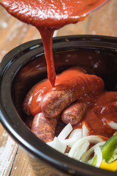 a person pouring sauce over hot dogs in a crock pot with onions, peppers and jalapenos