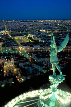 an aerial view of the city lights and buildings at night, including a statue in the foreground