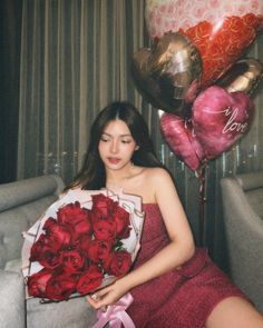 a woman sitting on a couch holding a bouquet of red roses and heart shaped balloons