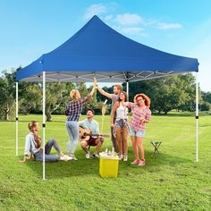 group of people standing under a blue tent in the grass with one woman holding her hand up