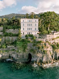 a large white house sitting on top of a cliff next to the ocean in front of a lush green hillside