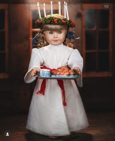 a doll dressed in white holding a plate with apples on it and candles behind her