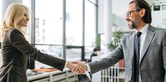 two business people shaking hands in an office setting with large windows on the wall behind them