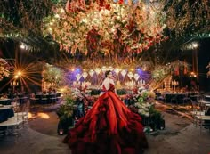 a woman in a long red dress standing under a chandelier filled with flowers