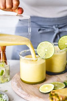 a person pouring a drink into two glasses with limes on the rim and pineapple slices around them