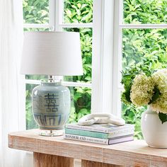 a table with a lamp, vase and books on it in front of a window