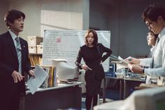 three people standing in an office with papers on the desk and one person holding a pen