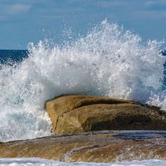 an ocean wave crashes on the rocks near the water's edge in front of a blue sky