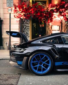 a black and blue car parked in front of a building with red flowers behind it