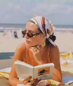 a woman reading a book on the beach while wearing sunglasses and a bandana hat