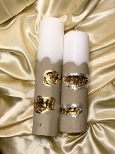 two white candles with gold designs on them sitting on a satin surface, next to each other