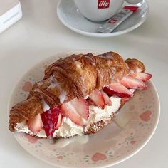 a croissant with cream and strawberries on it sitting on a plate next to a cup