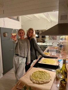 two women standing next to each other in a kitchen with pizzas on the counter