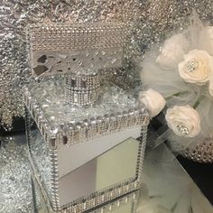 two white roses are sitting next to a silver box with diamonds on it and flowers in the background