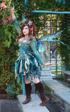 a woman dressed as a fairy standing in front of some bushes and flowers with her arms outstretched