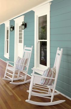 two white rocking chairs sitting on top of a wooden floor next to a blue house