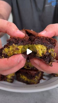 two hands holding up some brownies on a white plate with yellow and green frosting